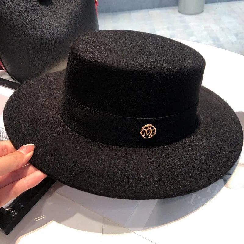Elegant black flat top fedora hat for women with gold emblem, on a table indoors